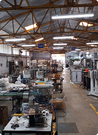 Wide-angle view of an industrial manufacturing facility with multiple packaging machines, assembly workstations, and production equipment arranged across a spacious factory floor.