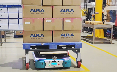 Autonomous mobile robot carrying a pallet loaded with stacked cardboard boxes in a manufacturing facility.