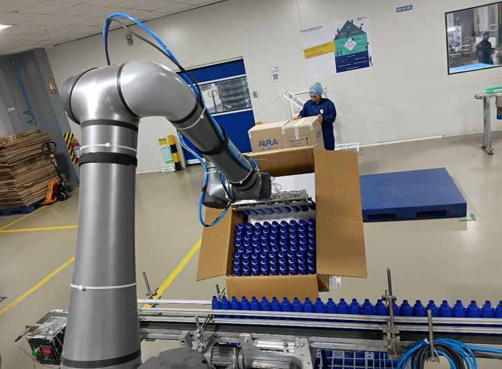 Robotic arm packing blue bottles into a cardboard box on an automated production line in a manufacturing facility.