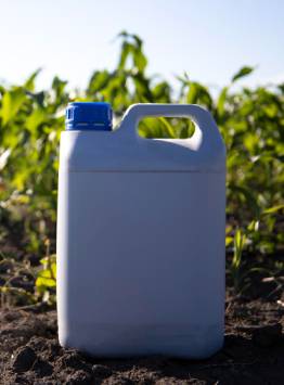 Large white plastic jerry can with an integrated carrying handle and blue screw cap, placed in an agricultural field. The container is commonly used for bulk agrochemical, fertilizer, and crop protection formulations.