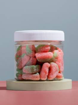 Transparent plastic jar with a white screw-on lid containing sugar-coated pink and green gummy candies, displayed on a pedestal against a soft pastel background.