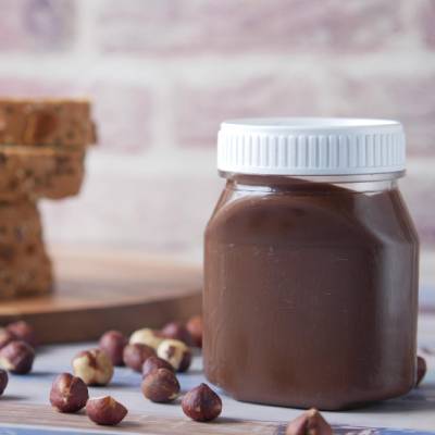 Clear plastic jar filled with chocolate spread or hazelnut paste, sealed with a white screw cap, placed on a wooden surface with scattered hazelnuts in the foreground.