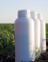 Three white HDPE plastic bottles with screw-cap closures placed in a cultivated agricultural field. The containers are commonly used for packaging agrochemical and crop protection products.