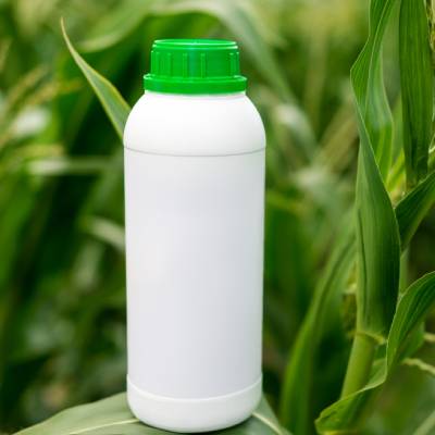 White HDPE plastic bottle with a green screw cap positioned among crop plants in an agricultural field. The container is commonly used for agrochemical and crop protection formulations.